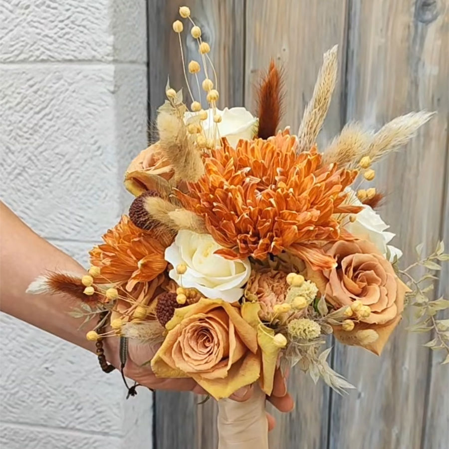 Brautstrauß mit getrockneten orangefarbenen Chrysanthemen, weißen Rosen, getrockneten Gräsern und weiteren Trockenblumen vor Holztür und weißer Wand