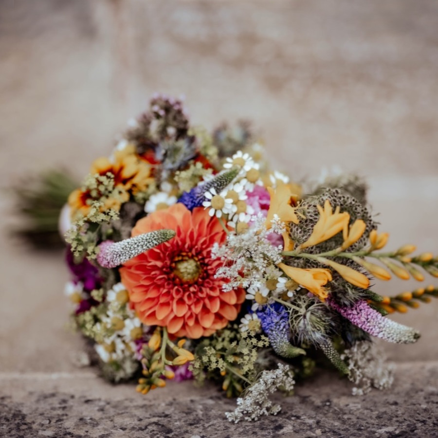 Bunter Blumenstrauß mit orangefarbener Blüte, kleinen weißen und lila Blüten sowie verschiedenen grünen Blättern auf Steinboden liegend