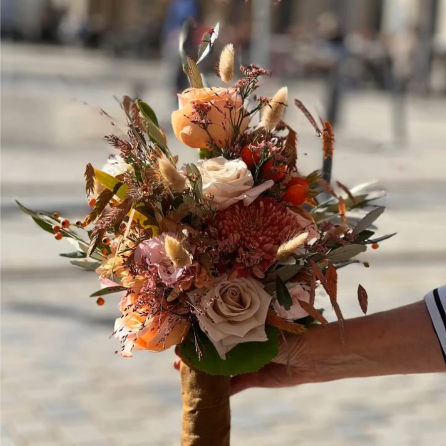 Brautstrauß mit Rosen, Chrysanthemen, Beeren und Trockenblumen in warmen Farbtönen, gehalten von einer Hand mit gestreiftem Ärmel
