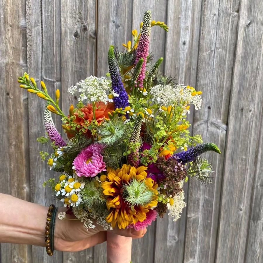 Bunte Blumenstrauß mit verschiedenen Blüten und grünen Zweigen vor einer Holzwand, gehalten von einer Hand mit Armband