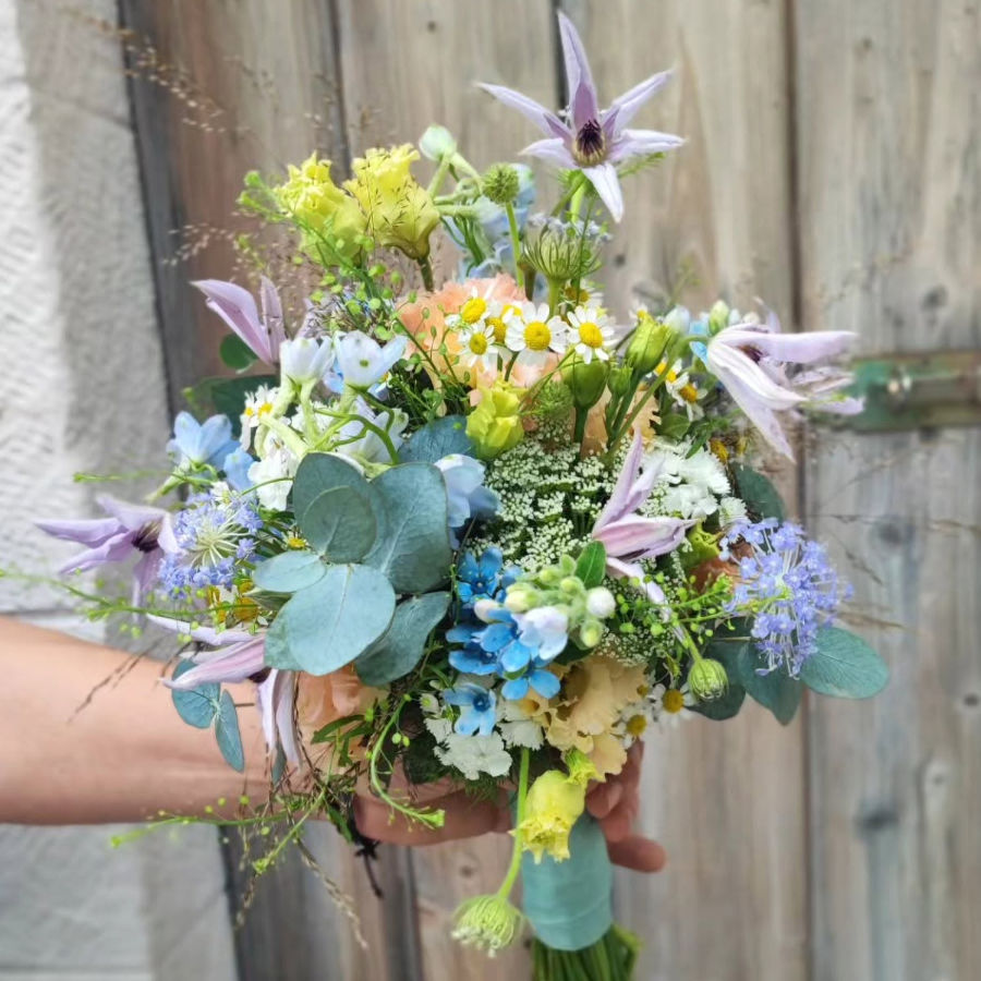 Bunter Blumenstrauß mit verschiedenen Blüten und grünen Blättern, gehalten von einer Hand vor einer Holzwand